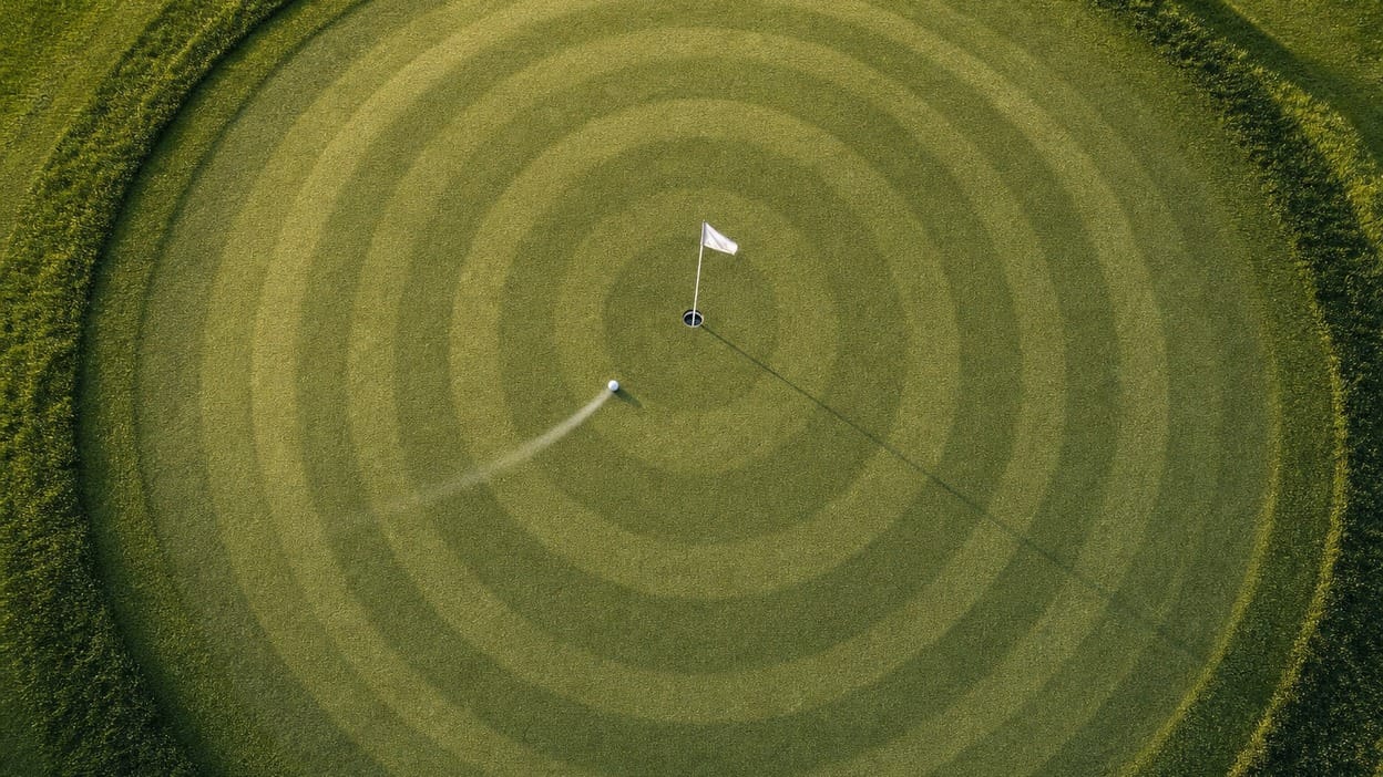 Aerial view of a putting green, concentric mowing-stripe rings around the flagstick, and a ball rolling toward the cup, showing a chip-shot landing-zone system.