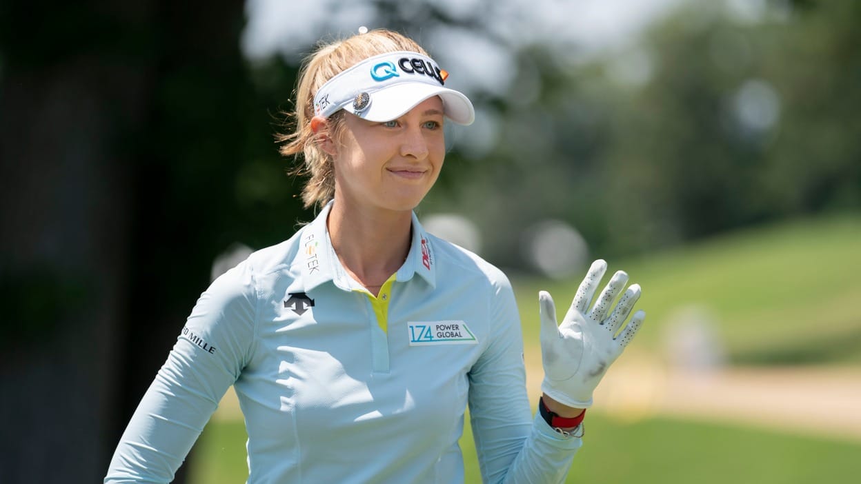 Nelly Korda smiles and waves to the gallery during an LPGA Tour event, wearing a light blue outfit, white visor, and golf glove.