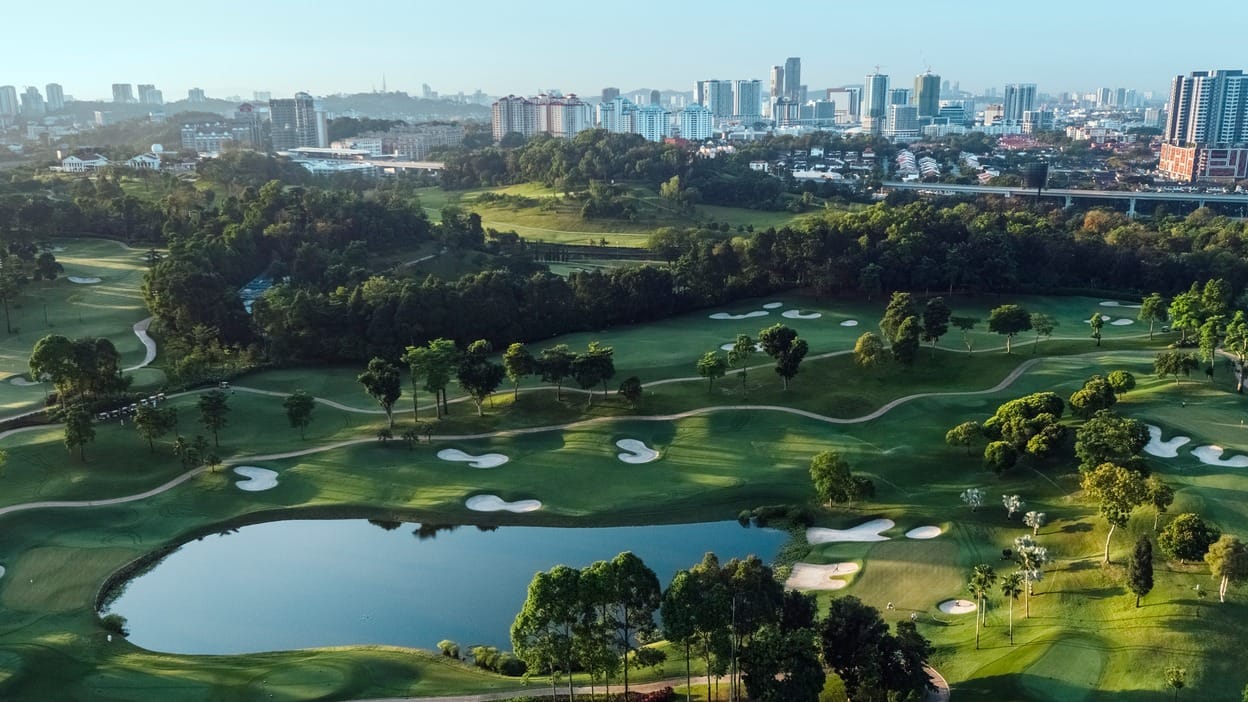 Aerial view of a golf course with bunkers, fairways and a lake surrounded by palm trees, with the hazy Kuala Lumpur skyline in the background, no players or signage visible.