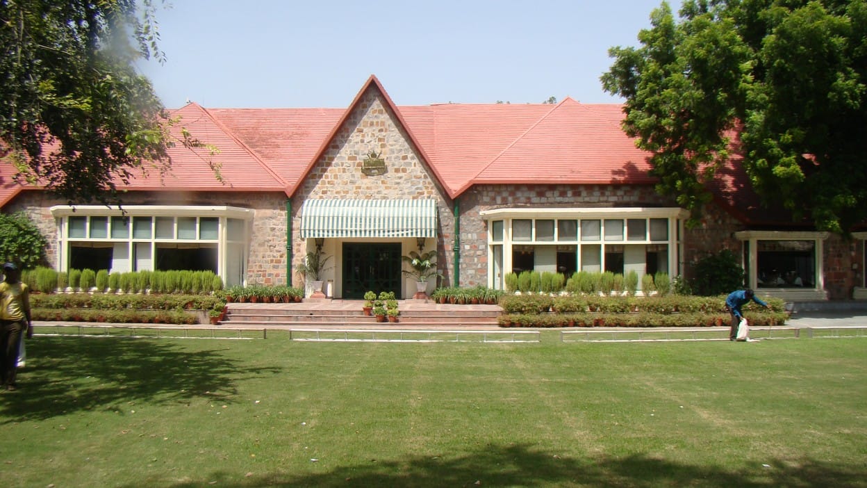 The red roofed stone clubhouse at Delhi Golf Club, with manicured lawn, hedges and trees in front under a clear sky.