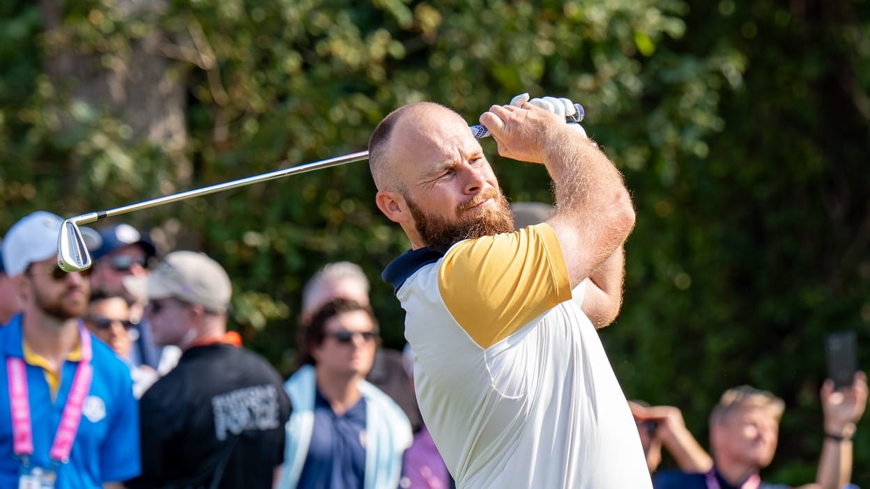 Tyrrell Hatton follows through on an iron shot in Europe's white and yellow 2025 Ryder Cup kit, gallery blurred behind him among trees.
