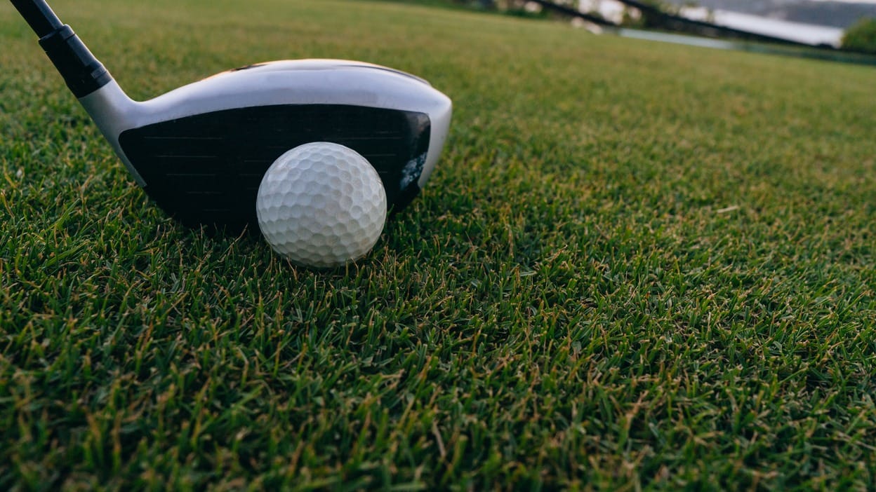 Close-up, ground-level view of a fairway wood clubhead addressing a golf ball on short, tightly mown fairway turf, no tee, with blurred grass and a distant fence in the background.