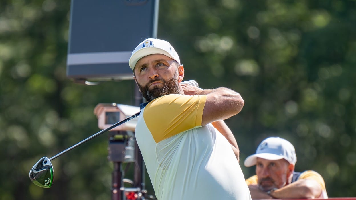 Jon Rahm finishing a driver swing in a white Ryder Cup cap and Europe team kit, a spectator in a cap blurred behind him. Dated 2025 Ryder Cup file photo, not a LIV Golf image.