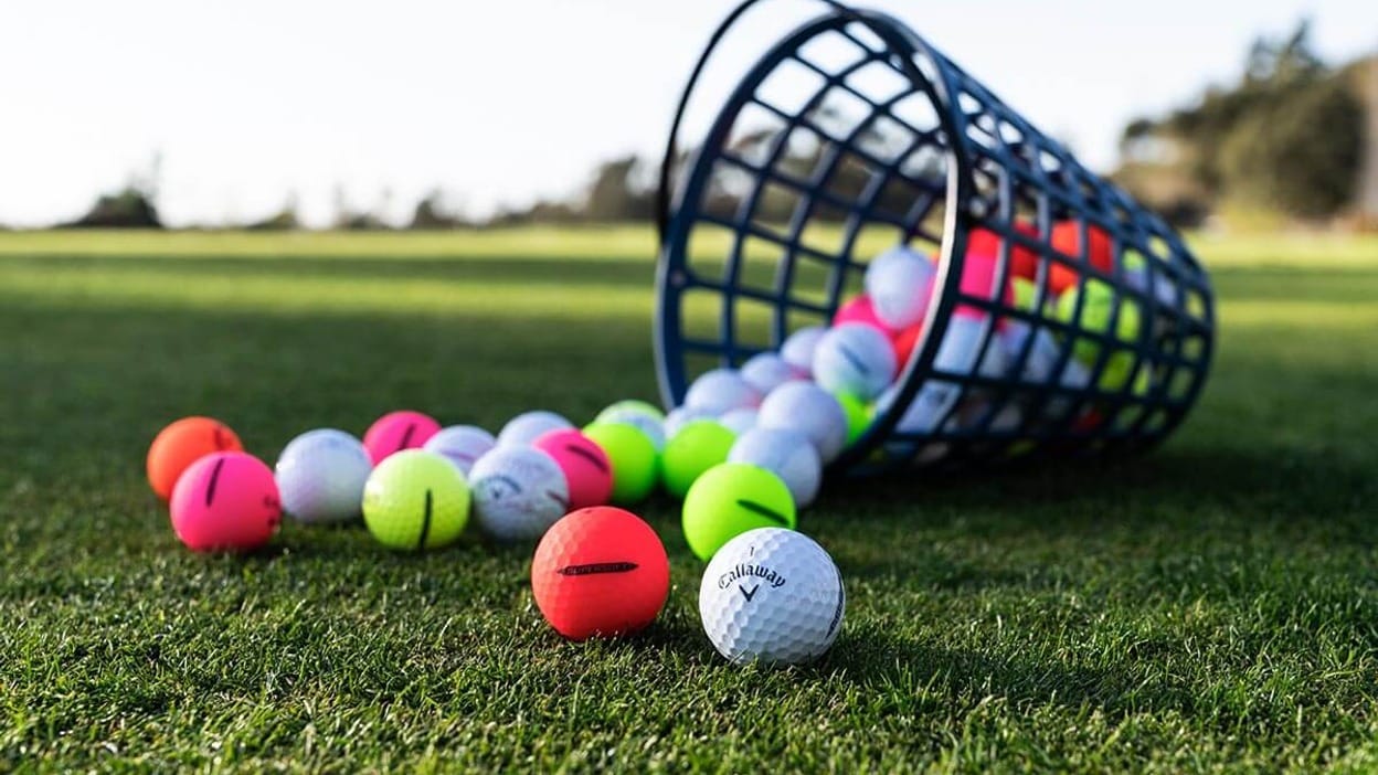 A Callaway Supersoft golf ball on grass in front of a tipped over bucket spilling colorful practice balls