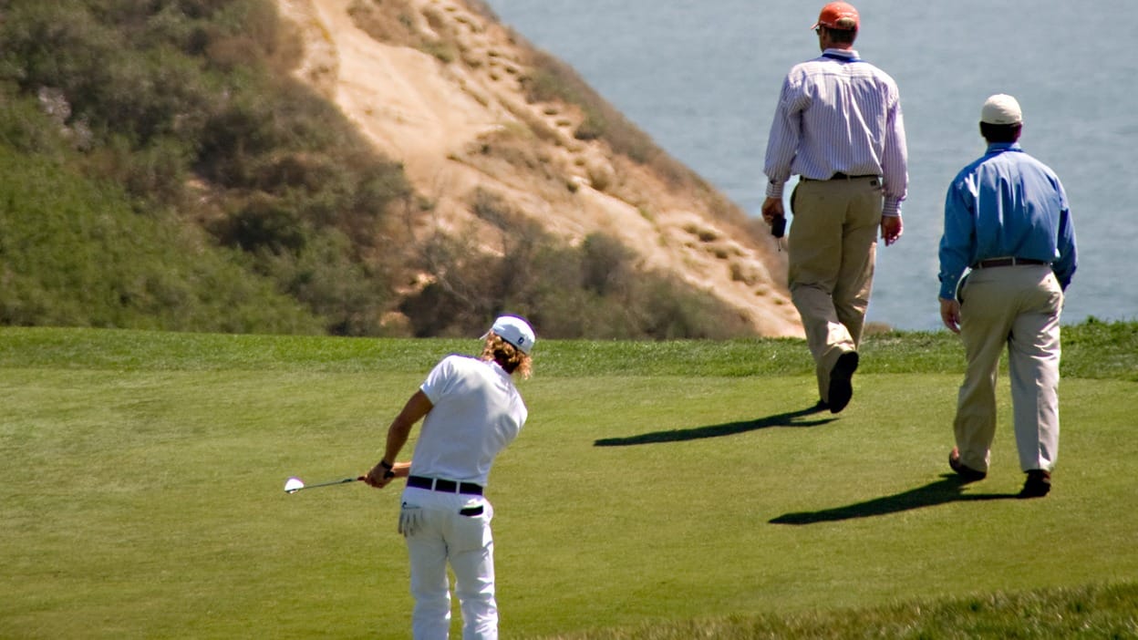 A golfer chipping on the 3rd hole at Torrey Pines South Course, with cliffs above the Pacific Ocean in the background.